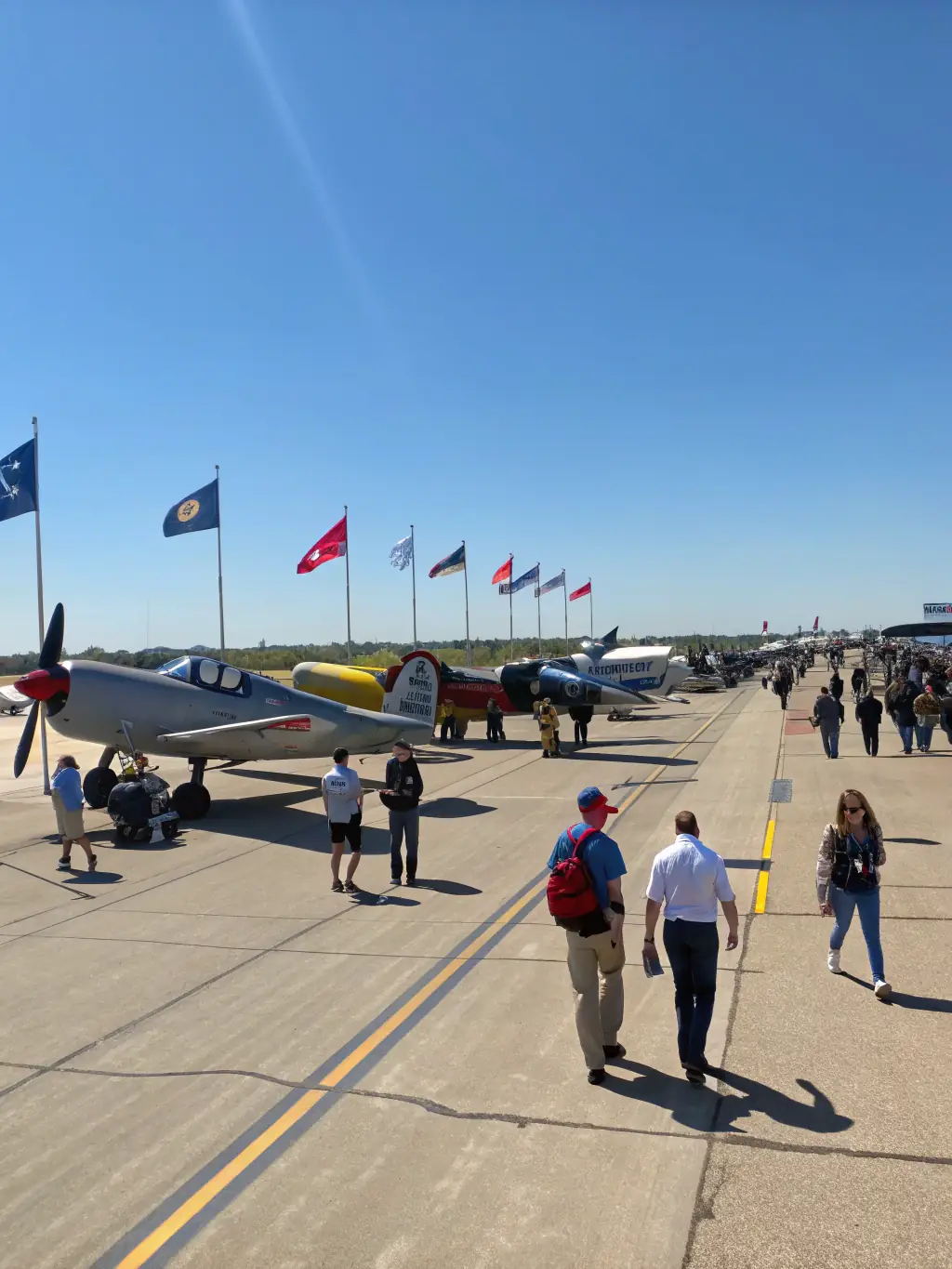 A festive scene of families and aviation enthusiasts attending an open day at the airfield, with static aircraft displays, food stands, and informational booths.